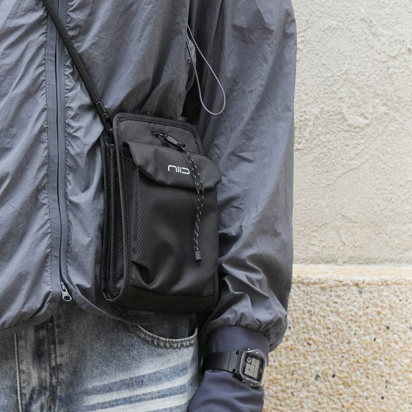 Urban lifestyle photo featuring black textured NIID mini crossbody phone bag with white logo and drawstring closure, worn over lightweight gray waterproof windbreaker jacket paired with faded blue jeans and square digital watch; neutral textured beige stone wall background minimalist street fashion product shot