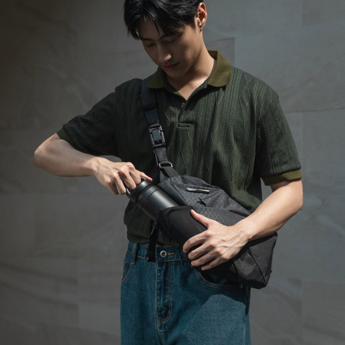 Waist-up lifestyle shot of young man in dark green textured polo shirt and blue jeans carrying dark gray grid NIID crossbody sling bag across chest; hands inserting black travel tumbler into side water bottle pocket, soft textured gray wall background urban daily carry product showcase