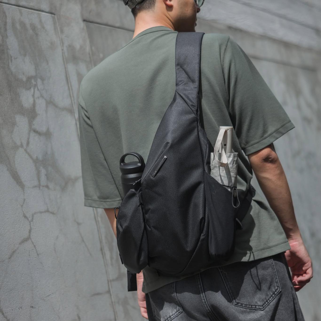 Back-view lifestyle photo of a man wearing muted green short-sleeve tee and gray jeans carrying dark gray NIID sling backpack with multi functional pockets; side water bottle holder, front slip pocket and small external pouch visible, textured cracked concrete wall background minimalist street travel product image