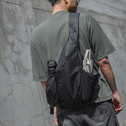 Back-view lifestyle photo of a man wearing muted green short-sleeve tee and gray jeans carrying dark gray NIID sling backpack with multi functional pockets; side water bottle holder, front slip pocket and small external pouch visible, textured cracked concrete wall background minimalist street travel product image