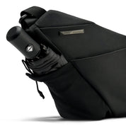 Close-up detail shot of Black Noir FINO V Ultra-slim Tech Sling crossbody bag side mesh slip pocket holding compact folding umbrella, engraved metal brand name plate on waterproof nylon exterior, showing multi-purpose outdoor storage feature on clean white studio background