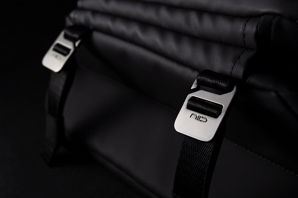 Dark background close-up of two engraved NIID silver metal buckles on meteorite black NIID R1 Radiant Urban Sling crossbody bag, thick woven nylon shoulder straps and folded waterproof nylon fabric, premium hardware detail shot highlighting durable adjustable fasteners for daily carry