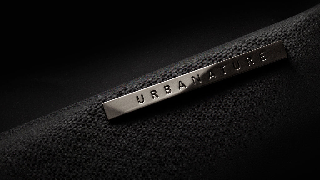 Extreme close-up shot of engraved URBANATURE metal brand plate on meteorite black NIID R1 Radiant Urban Sling crossbody bag, sleek matte metallic logo detail on smooth waterproof nylon fabric with soft shadow dark background