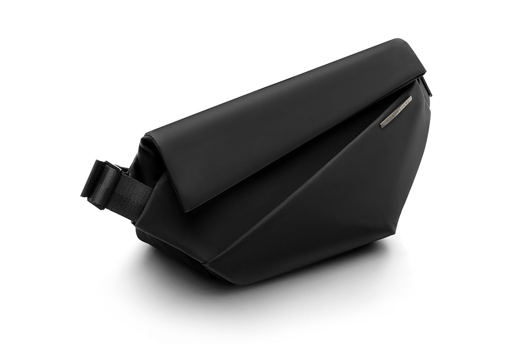 Side angle shot of meteorite black NIID R1 Radiant Urban Sling crossbody bag, waterproof nylon unisex anti-theft chest pack with adjustable shoulder strap and engraved metal brand plate, minimalist fold-over design on clean white background for daily commuting and travel