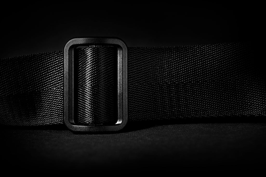 Monochrome dark mood close-up of textured plastic slider adjuster on thick woven nylon shoulder strap for NIID R1 Radiant Urban Sling crossbody bag, tight weave fabric detail, minimalist black background showcasing durable adjustable strap hardware
