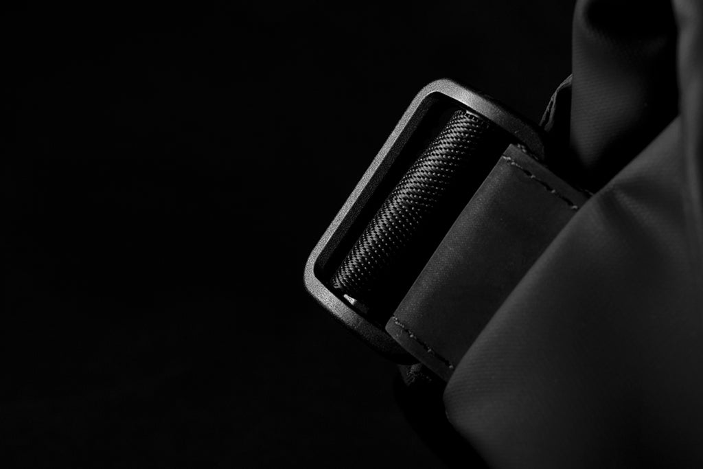 Dark moody close-up of textured plastic shoulder strap adjuster buckle on meteorite black NIID R1 Radiant Urban Sling crossbody bag, fine stitching on waterproof nylon strap, minimalist dark background highlighting hardware craftsmanship