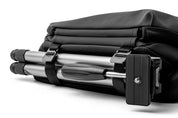 Close-up detail of meteorite black NIID R1 Radiant Urban Sling crossbody bag built-in tripod storage sleeve, holding foldable silver camera tripod with branded nylon fixing straps, waterproof nylon photography sling bag accessory compartment shot on plain white background
