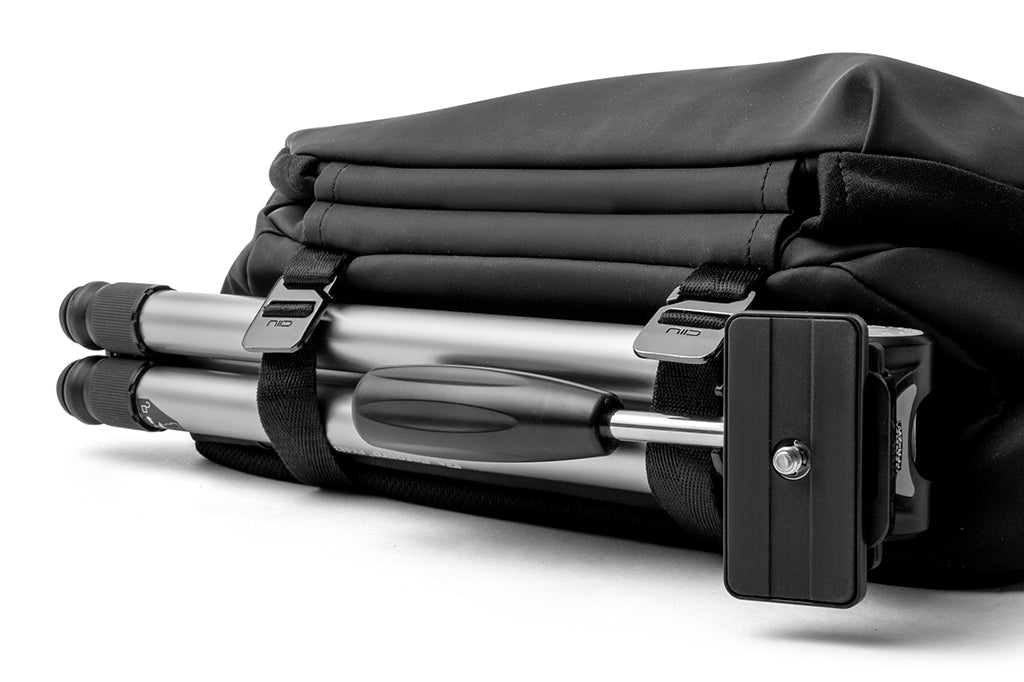 Close-up detail of meteorite black NIID R1 Radiant Urban Sling crossbody bag built-in tripod storage sleeve, holding foldable silver camera tripod with branded nylon fixing straps, waterproof nylon photography sling bag accessory compartment shot on plain white background