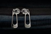 Close-up shot of two engraved NIID metal zipper pulls on textured indigo woven bag fabric, premium double zipper hardware detail showcase