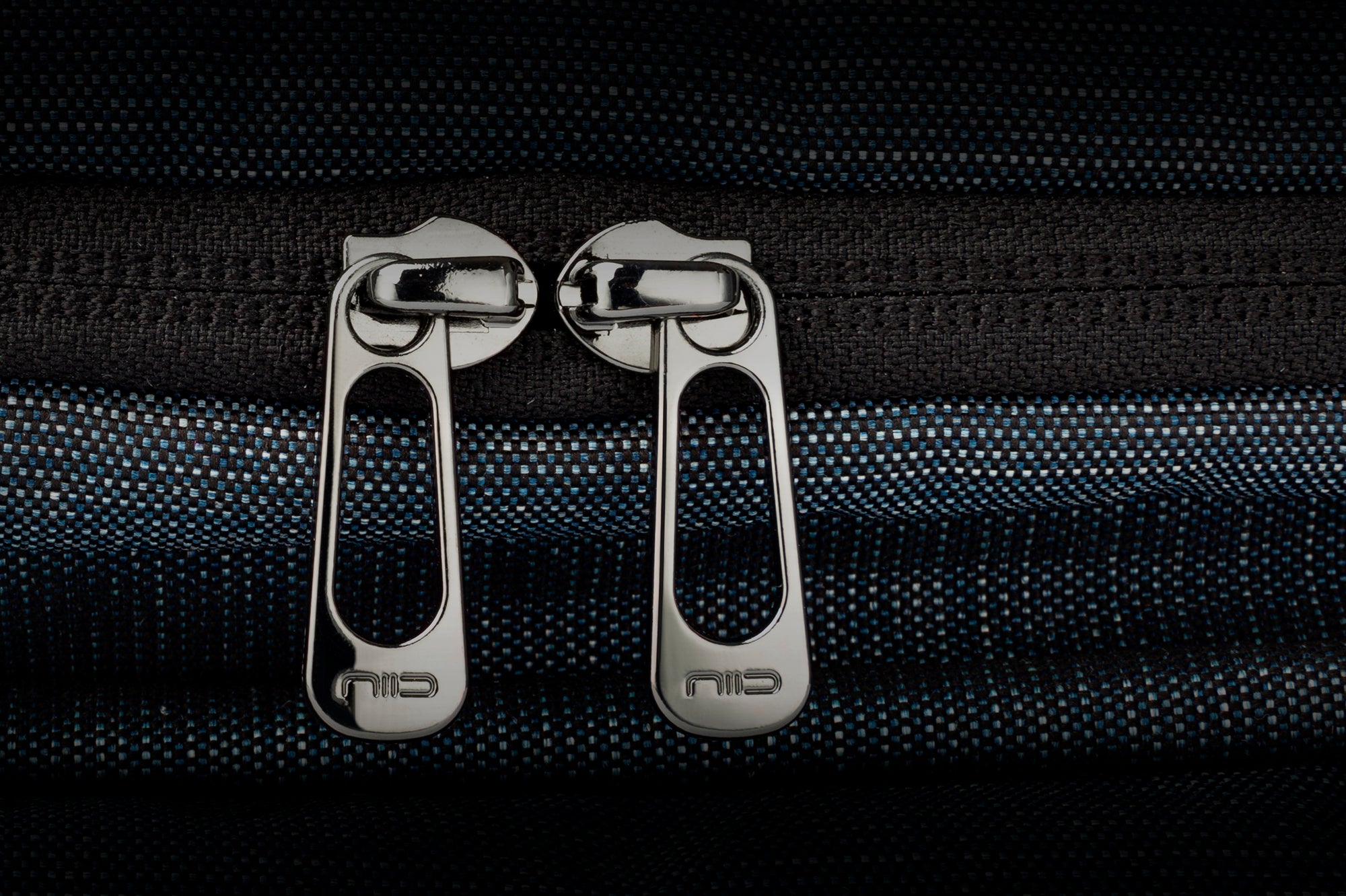 Close-up shot of two engraved NIID metal zipper pulls on textured indigo woven bag fabric, premium double zipper hardware detail showcase