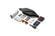 Flat lay shot of black NIID RO Radiant crossbody chest bag arranged with daily travel essentials including smartphone, passport, sunglasses, water bottle, wallet, car keys, earbuds and bank cards, showcasing large storage capacity