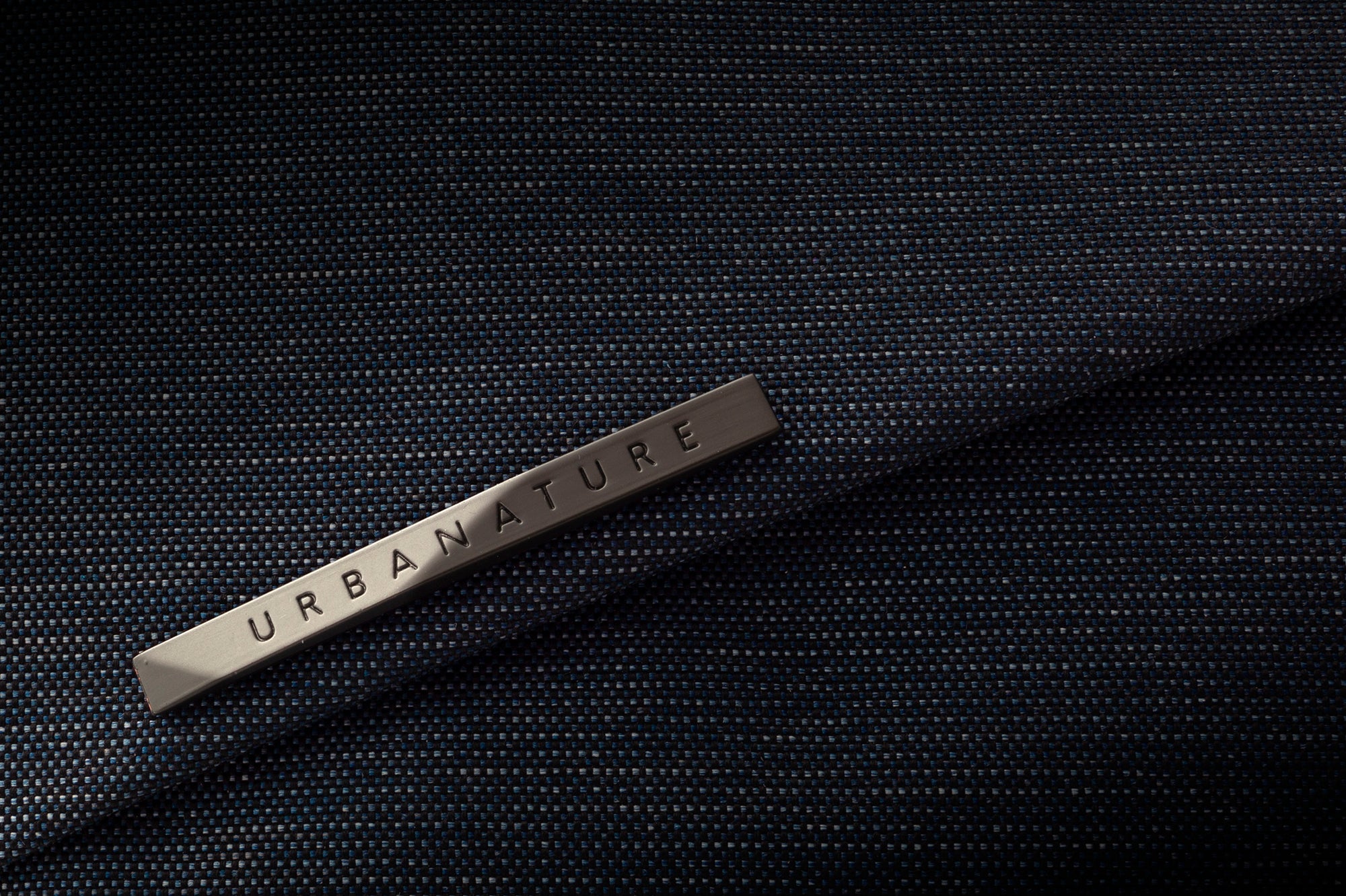 Close-up detail shot of metallic engraved URBANATURE brand tag laid on textured dark indigo woven waterproof bag fabric, premium material texture showcase