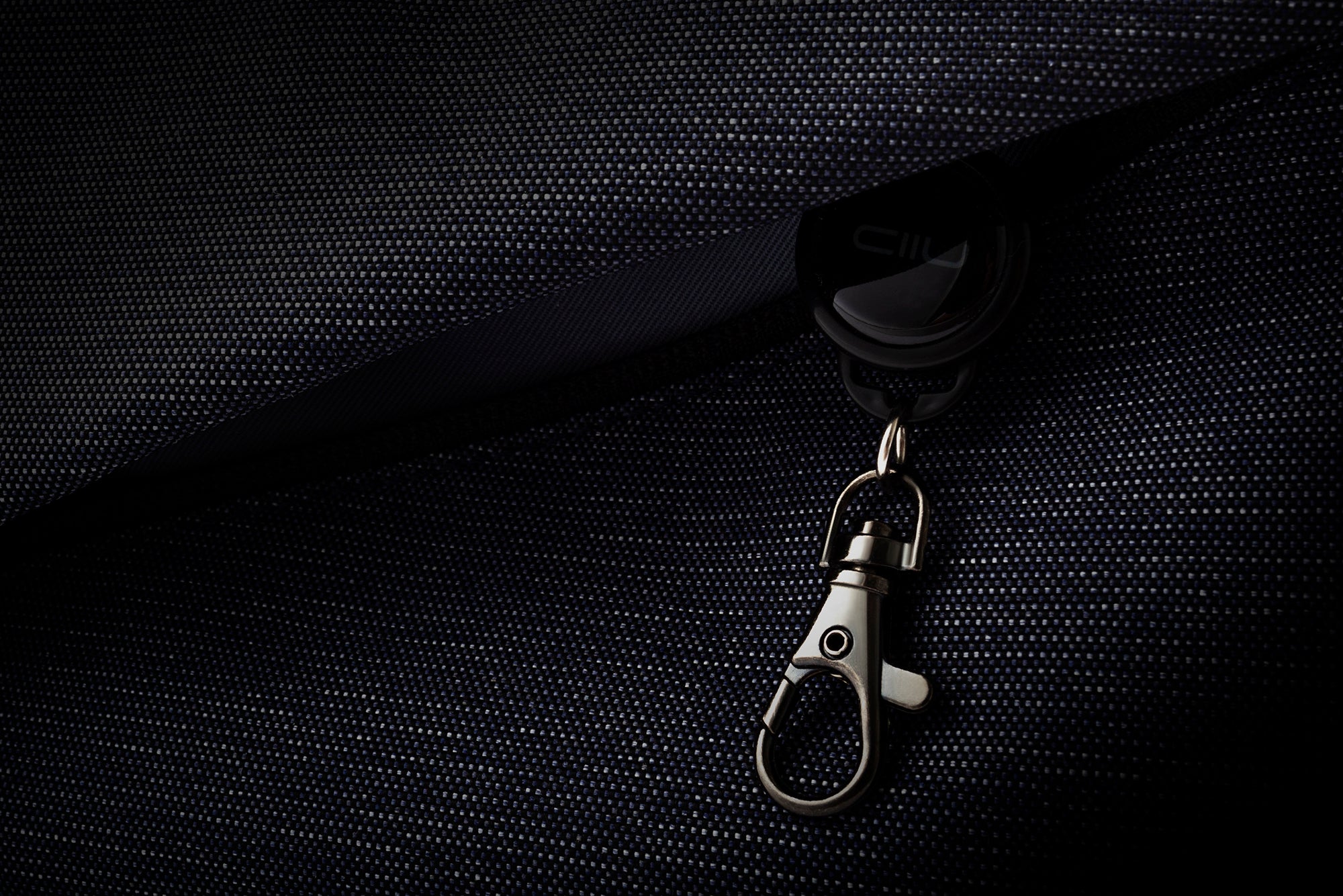 Dark mood close-up of metallic lobster claw hook accessory attached to indigo textured woven bag fabric, premium hardware detail shot