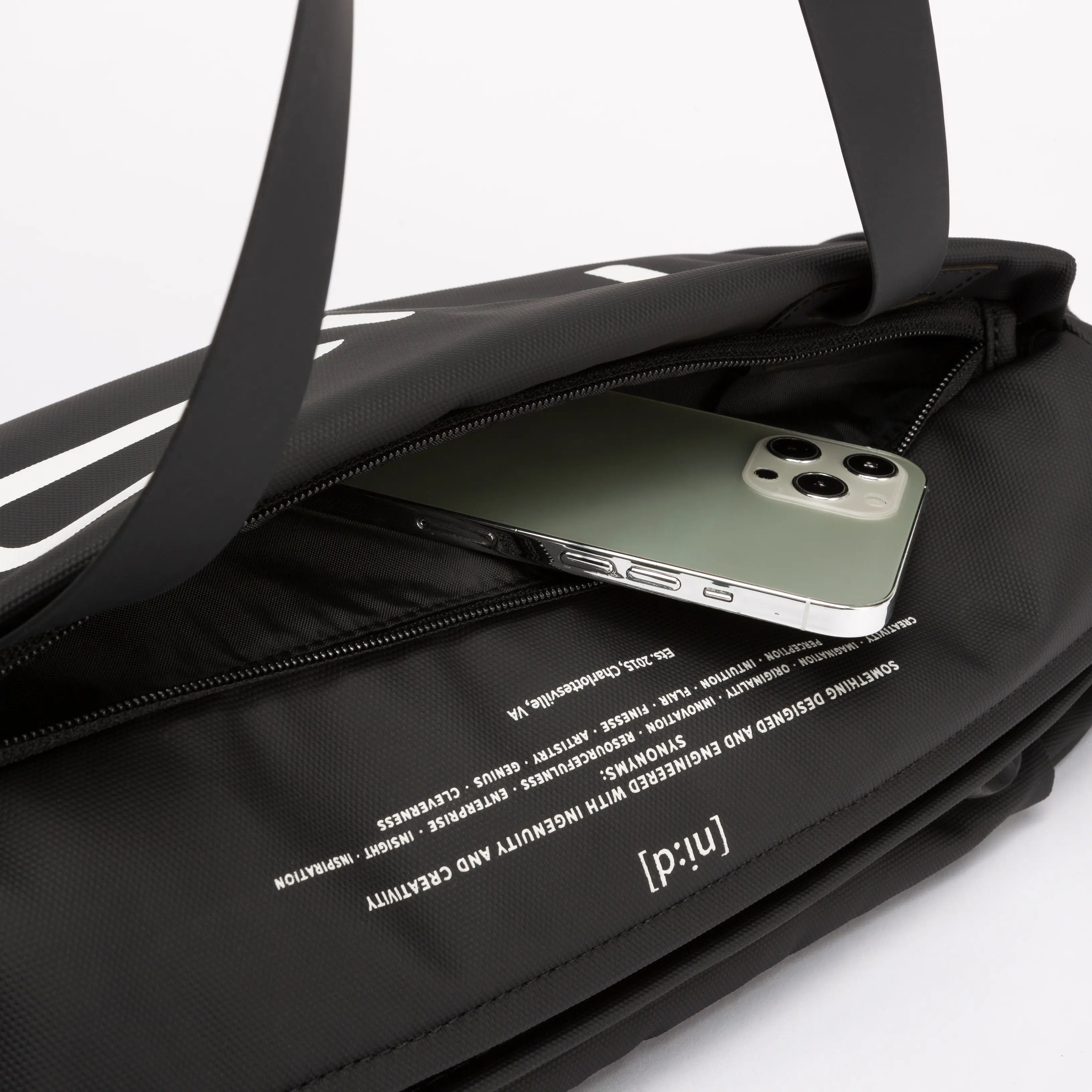 Close-up detail shot of black NIID S6 Hybrid Sling crossbody bag, front zipper pocket holding smartphone, printed NIID brand text and slogan fabric lining, dual top handles, plain white studio background