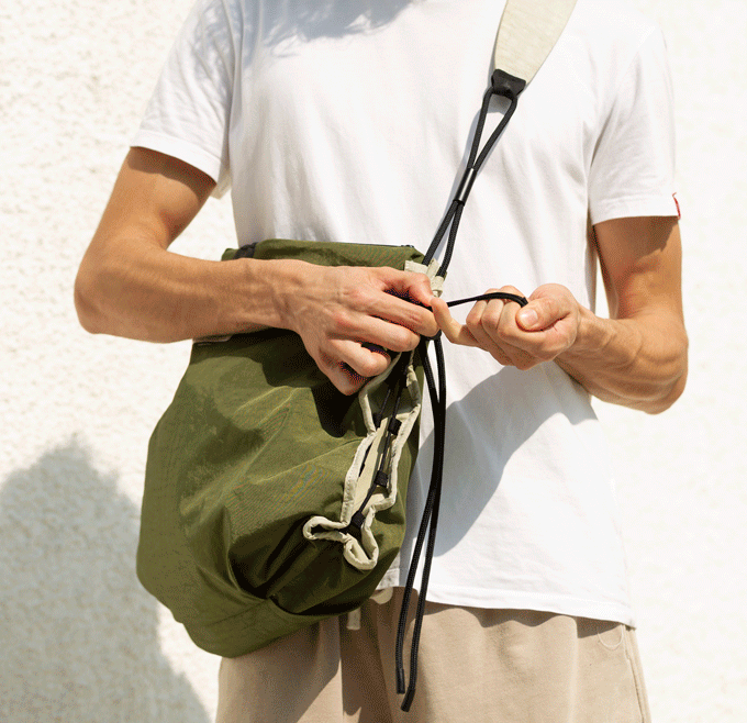 Close-up lifestyle detail shot of Khaki/Olive NIID ST@TEMENT S7 Tote Bag, male hands pulling the built-in drawstring closure, olive green lightweight fabric, matching drawstring toggle, adjustable mixed-texture shoulder strap detail, plain white textured wall background, showcase of the bag’s cinch feature