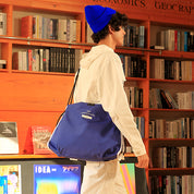 Square bookstore lifestyle shot of White/Navy NIID ST@TEMENT S7 Tote crossbody bag, male model with bright blue beanie and all-white outfit, wooden bookshelf background, navy blue fabric bag with white trim and front woven label, casual indoor daily carry showcase