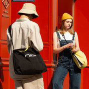 Square lifestyle street shot of NIID ST@TEMENT S7 Tote Bag, two models carrying Black and Buttercup colorway crossbody tote bags, bright red wall urban outdoor scene, branded yellow zipper tape and NIID front logo, casual daily commute outfit showcase