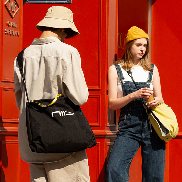 Square lifestyle street shot of NIID ST@TEMENT S7 Tote Bag, two models carrying Black and Buttercup colorway crossbody tote bags, bright red wall urban outdoor scene, branded yellow zipper tape and NIID front logo, casual daily commute outfit showcase