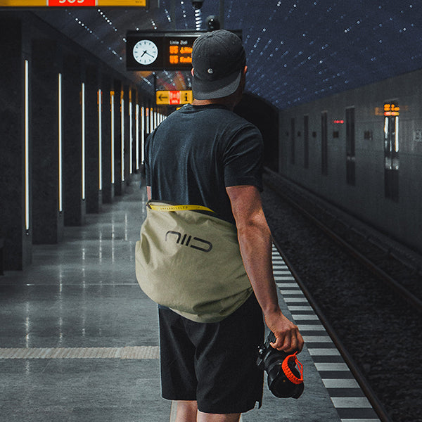 Olive colorway, male model viewed from back wearing the crossbody tote, bright yellow branded zipper tape, NIID front logo, urban transit commute scene, model holding camera with orange lens cap