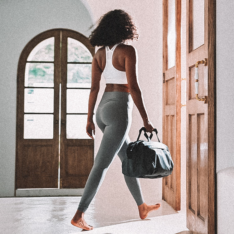 Woman in white sports bra and gray leggings carrying NIID CACHE Hybrid Tech Sling in Dark by top handle, gym lifestyle shot.