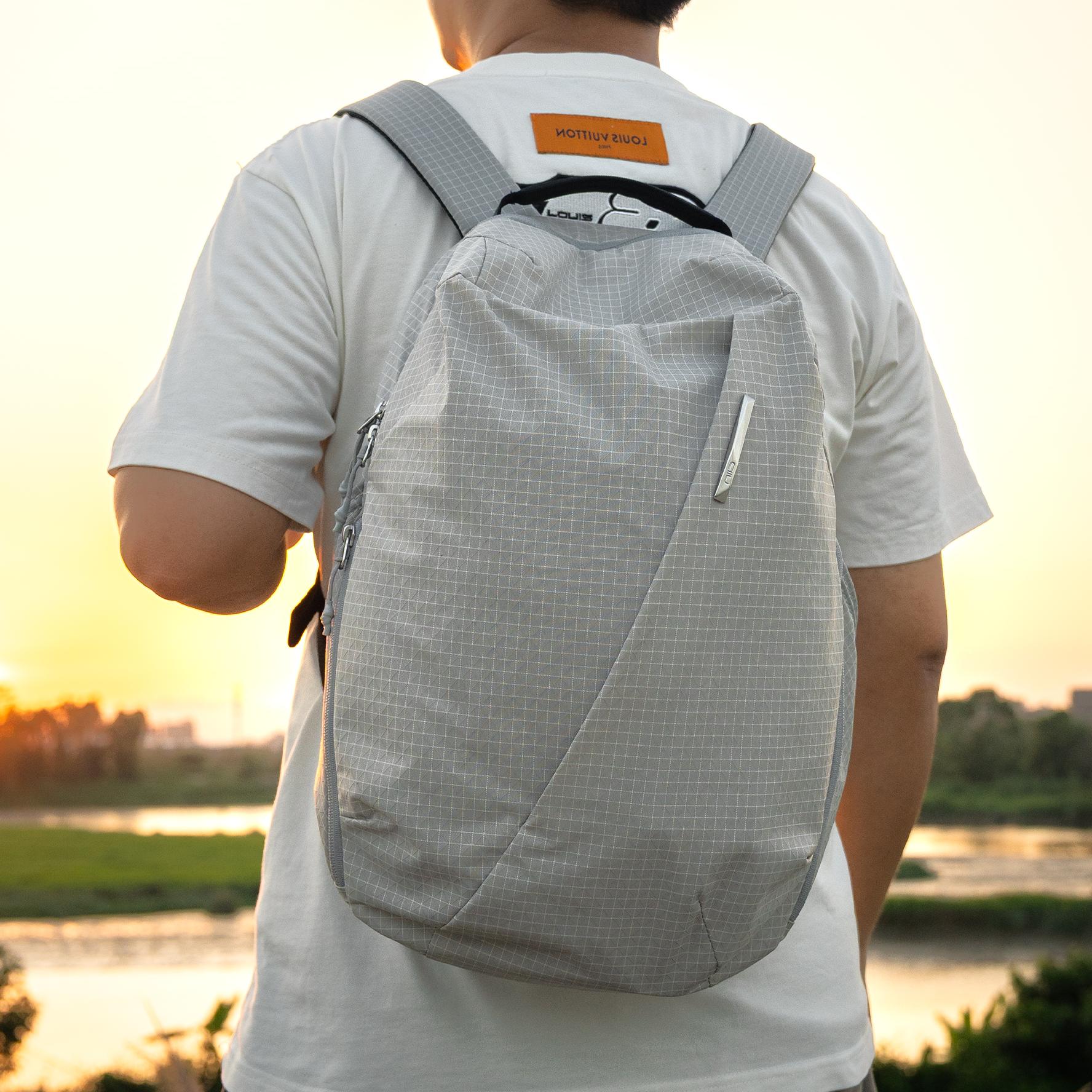 Back view of male model carrying NIID NEO Nomad 19L Backpack‑N3 glacier gray grid fabric at sunset riverside scene, lightweight water‑resistant daypack for urban travel and daily commute.