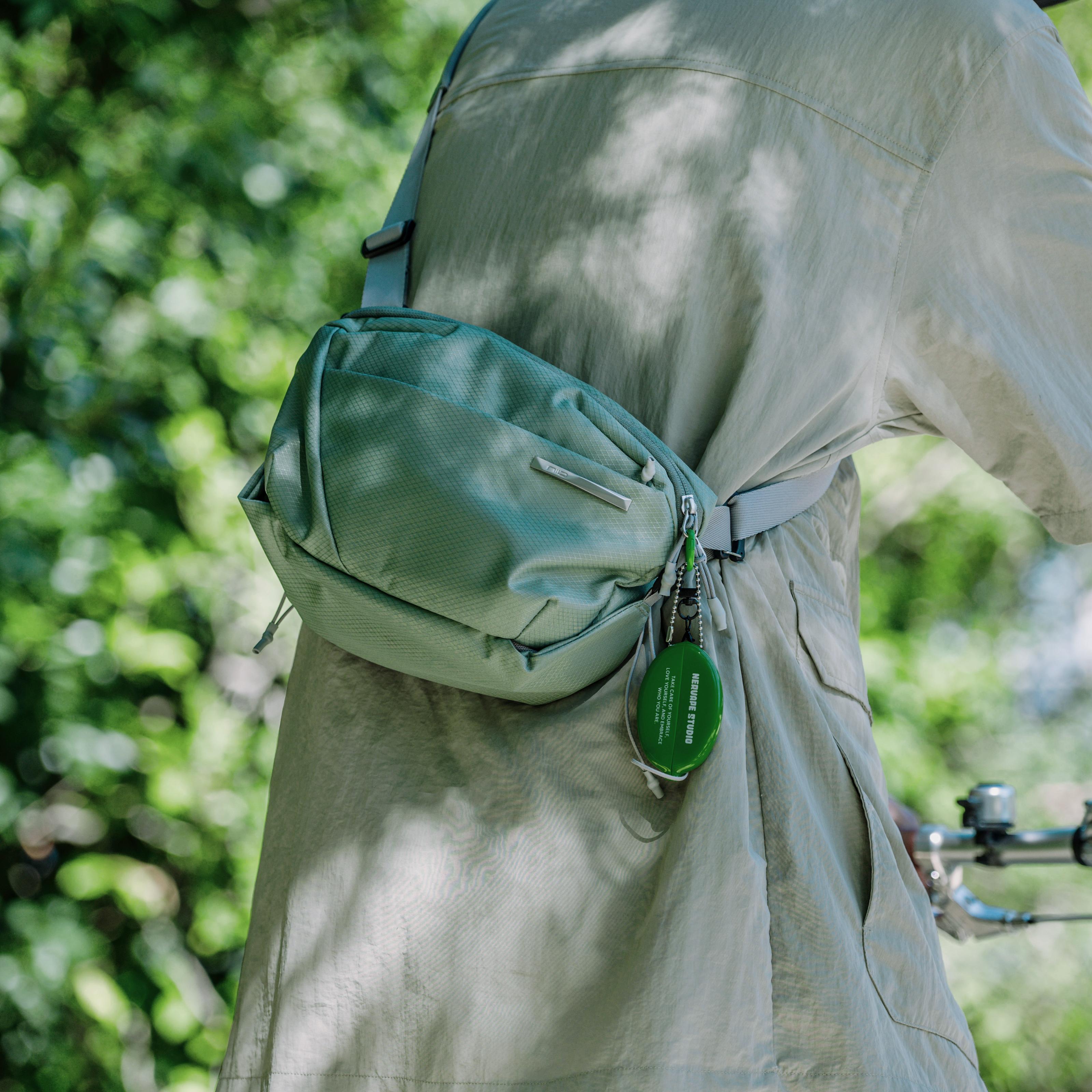 NIID NEO Nomad 4L Sling Bag‑N1 sage green diamond grid sling bag side worn close‑up, accessory keychain detail, blurred green foliage background, outdoor casual EDC crossbody bag