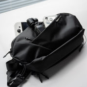 Studio shot of NIID NEO Nomad 5L Sling Bag‑N1L black textured crossbody sling bag, partially filled with smartphone, action camera and daily EDC gear on clean light‑gray table surface.