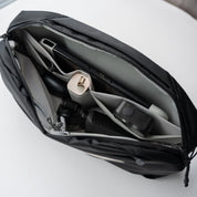 Studio shot of NIID NEO Nomad 5L Sling Bag‑N1L black textured crossbody sling bag, partially filled with smartphone, action camera and daily EDC gear on clean light‑gray table surface.