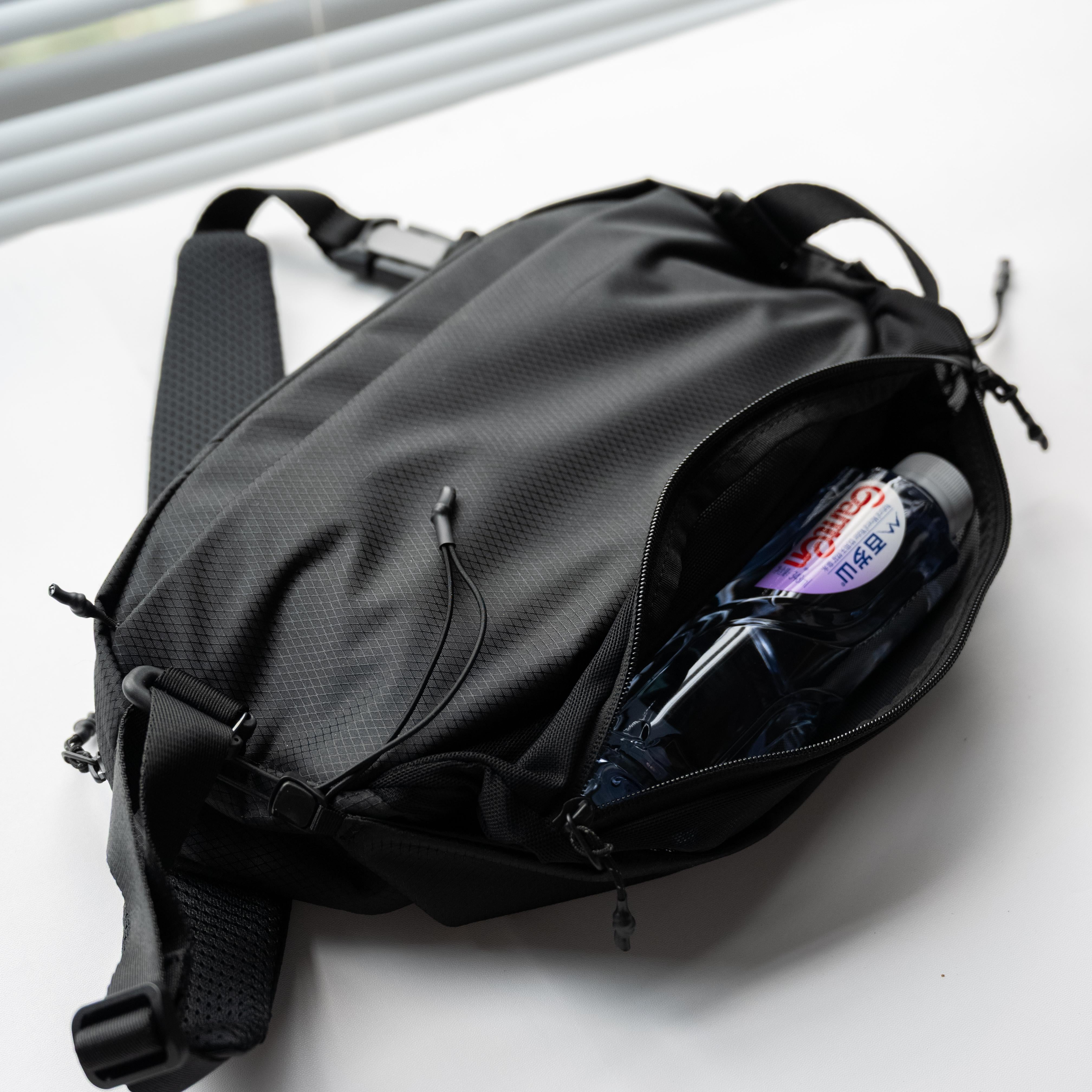 Side view of NIID NEO Nomad 5L Sling Bag‑N1L black, expandable mesh side pocket holding plastic water bottle, demonstrate flexible storage capacity for outdoor commute.