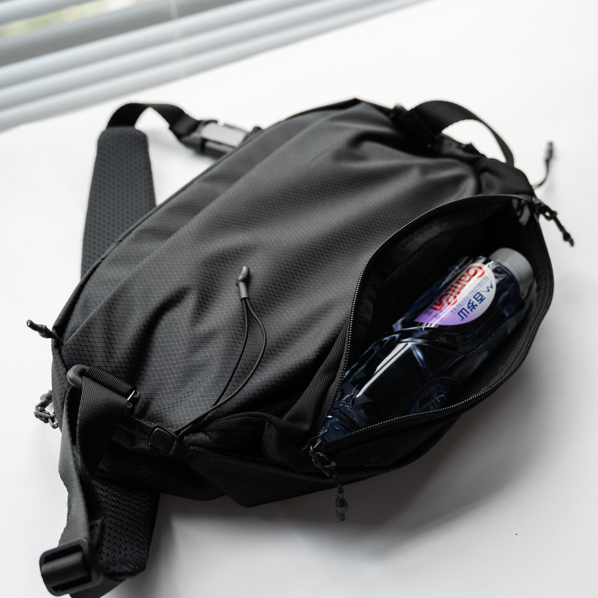Side view of NIID NEO Nomad 5L Sling Bag‑N1L black, expandable mesh side pocket holding plastic water bottle, demonstrate flexible storage capacity for outdoor commute.