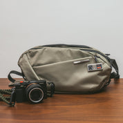 Flat‑lay table shot of NIID NEO Nomad 5L Sling Bag‑N1L pearly gray textured crossbody bag, placed beside compact Sony camera on wooden table, everyday EDC travel gear display.