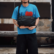 Street lifestyle photo of a man holding NIID NEO Nomad 9L black sling crossbody bag, wearing blue heart print tee and black loose pants, industrial weathered wall background, unisex everyday chest bag outdoor shooting