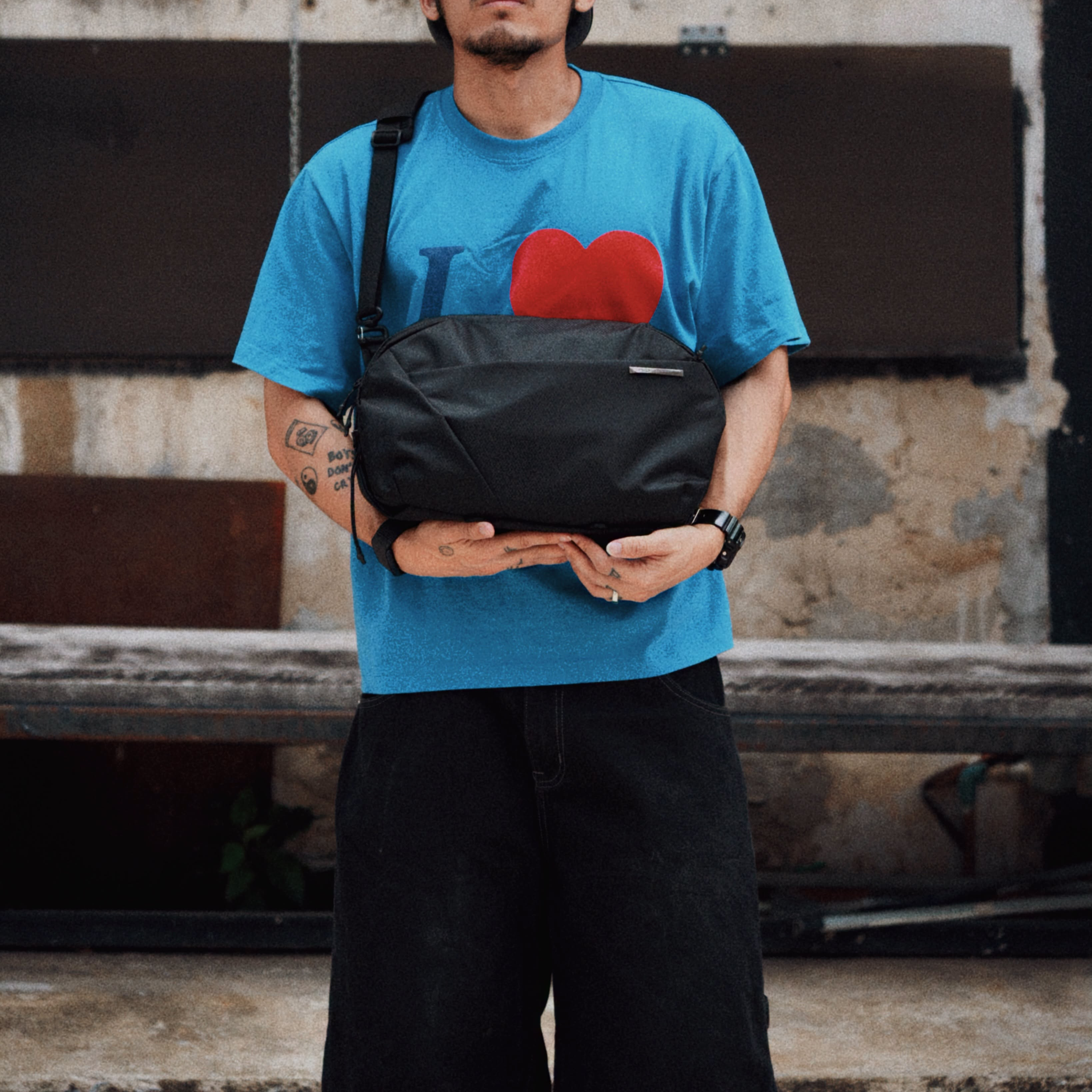 Street lifestyle photo of a man holding NIID NEO Nomad 9L black sling crossbody bag, wearing blue heart print tee and black loose pants, industrial weathered wall background, unisex everyday chest bag outdoor shooting