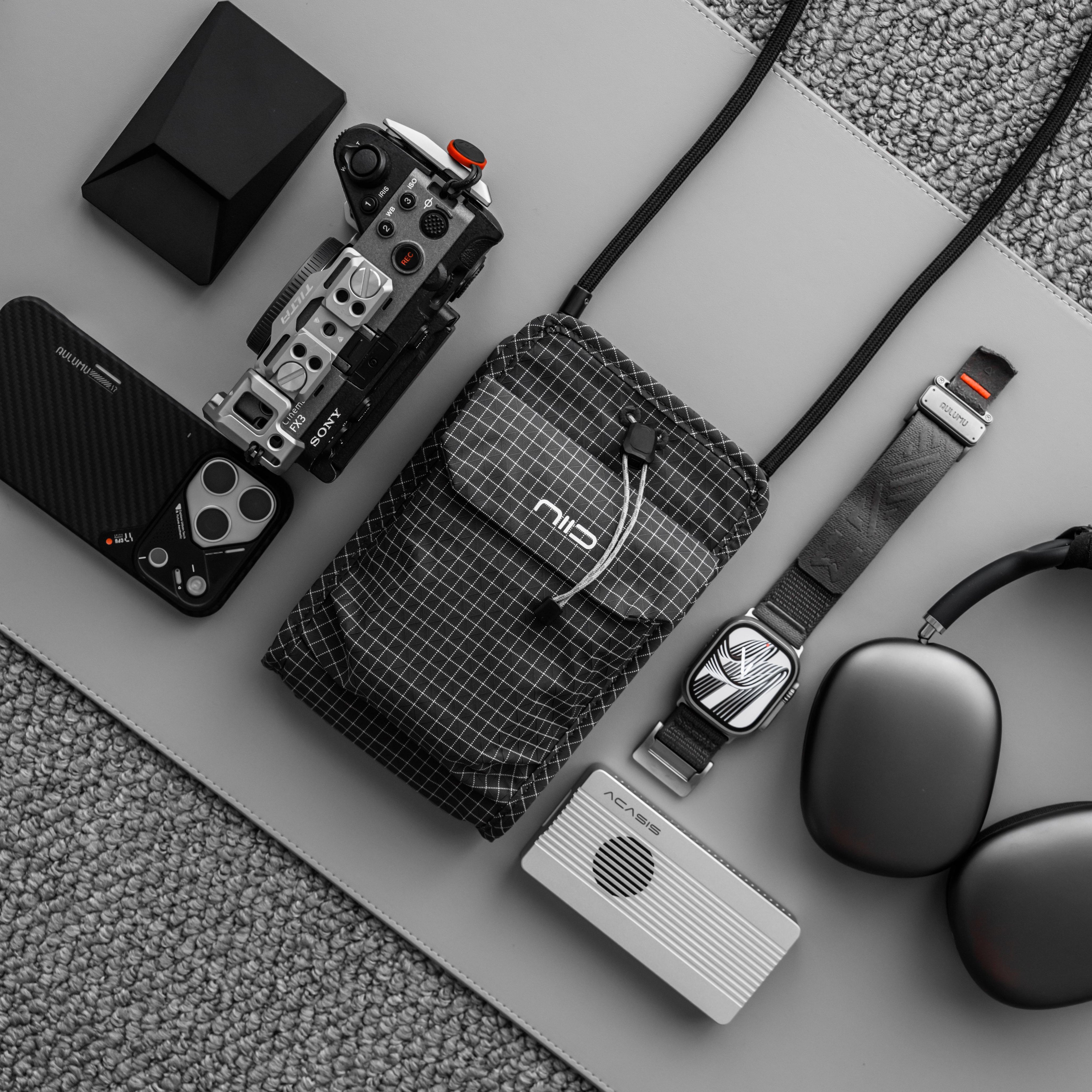 Flat lay of black diamond grid black NIID NEO Nomad Pouch with camera, phone, watch and headphones, lightweight crossbody pouch for tech gadgets