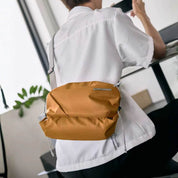 Back view of a person wearing plain white short sleeve shirt, carrying NIID NEO Nomad amber orange waterproof crossbody sling bag with silver metal brand plaque and light gray adjustable shoulder strap, bright minimalist indoor room with green plant decor, casual daily commute fashion shot, soft natural daylight, no watermarks