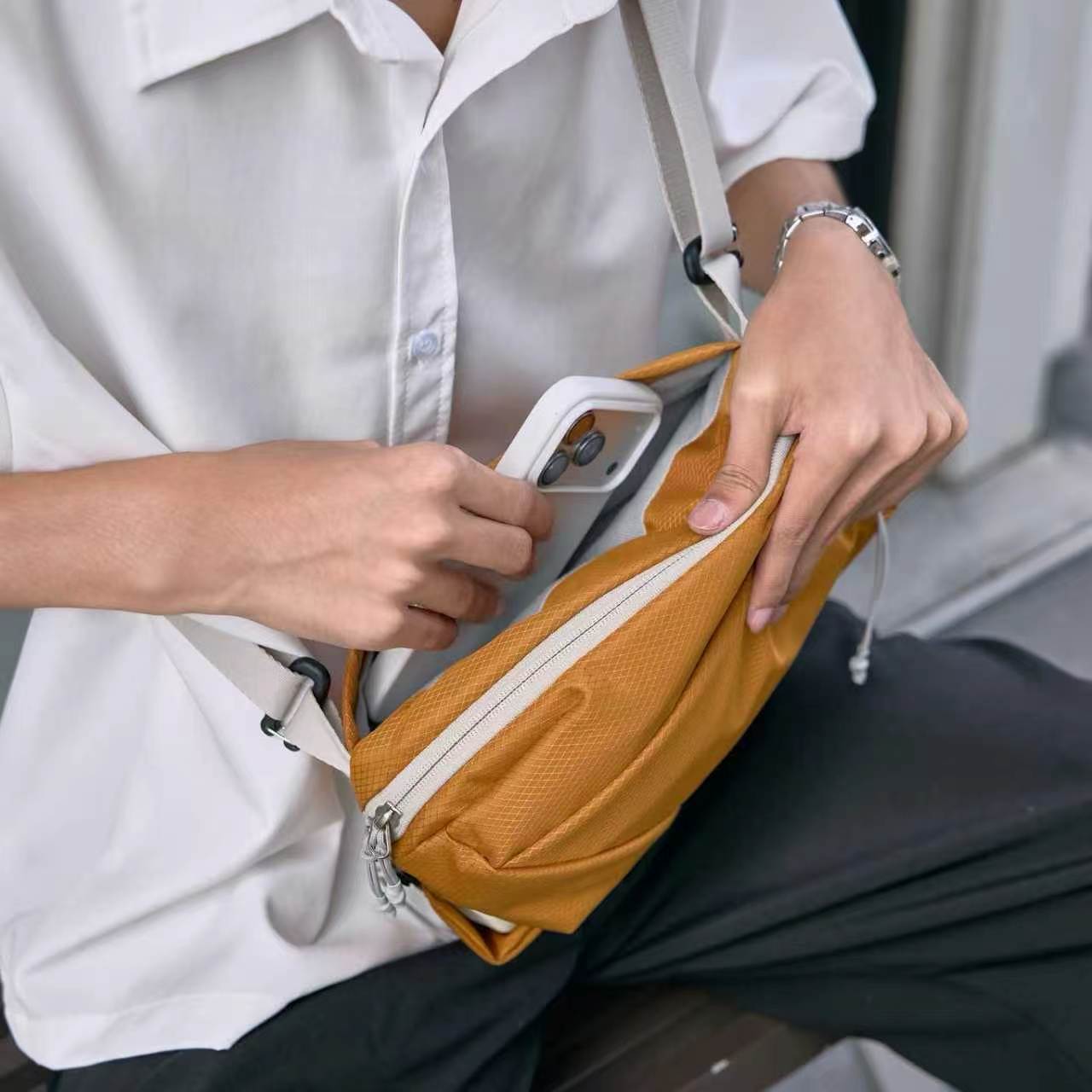 Close-up lifestyle shot of NIID NEO Nomad amber orange waterproof crossbody sling bag, person in white button-down shirt demonstrating front zipper pocket storage by placing a white smartphone inside, light gray adjustable shoulder strap, textured diamond grid fabric, casual outdoor bench scene, soft natural light, no watermarks