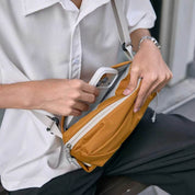 Close-up lifestyle shot of NIID NEO Nomad amber orange waterproof crossbody sling bag, person in white button-down shirt demonstrating front zipper pocket storage by placing a white smartphone inside, light gray adjustable shoulder strap, textured diamond grid fabric, casual outdoor bench scene, soft natural light, no watermarks