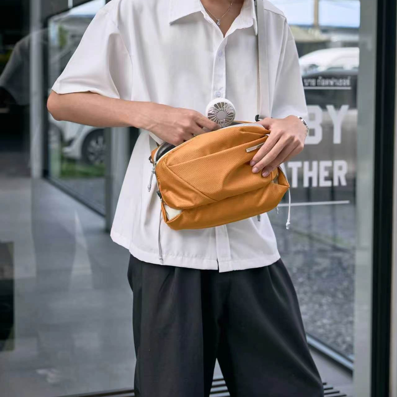 Male model in white short sleeve shirt and black casual trousers carrying NIID NEO Nomad amber orange waterproof crossbody sling bag at waist, hands opening front zipper pocket with mini fan inside, modern glass storefront background, urban streetwear photography, soft natural daylight, no watermarks
