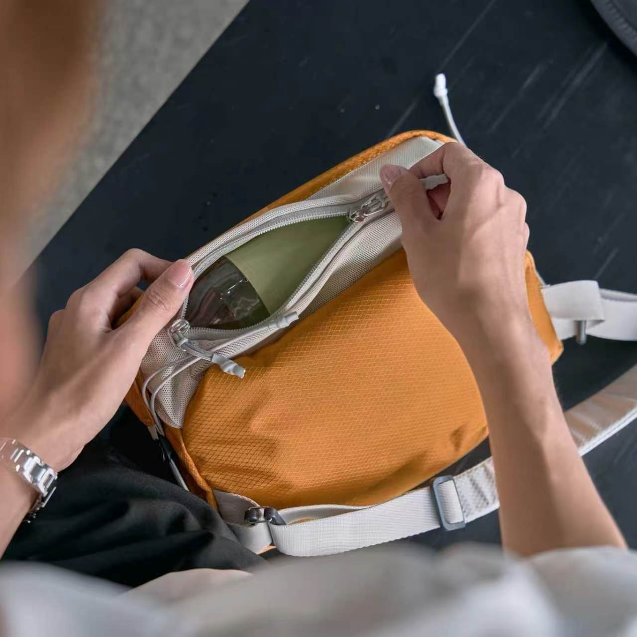 Top-down close-up shot of hands unzipping NIID NEO Nomad amber orange crossbody sling bag, showing spacious interior compartment with small earbuds stored inside, diamond grid waterproof fabric, light gray lining and adjustable shoulder strap, dark wooden table surface background, clean product detail photo without watermarks