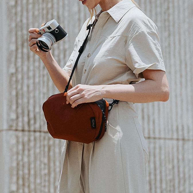 Woman in beige outfit carrying Venetian Red Tech Pouch as crossbody, holding Leica camera, street lifestyle shot.