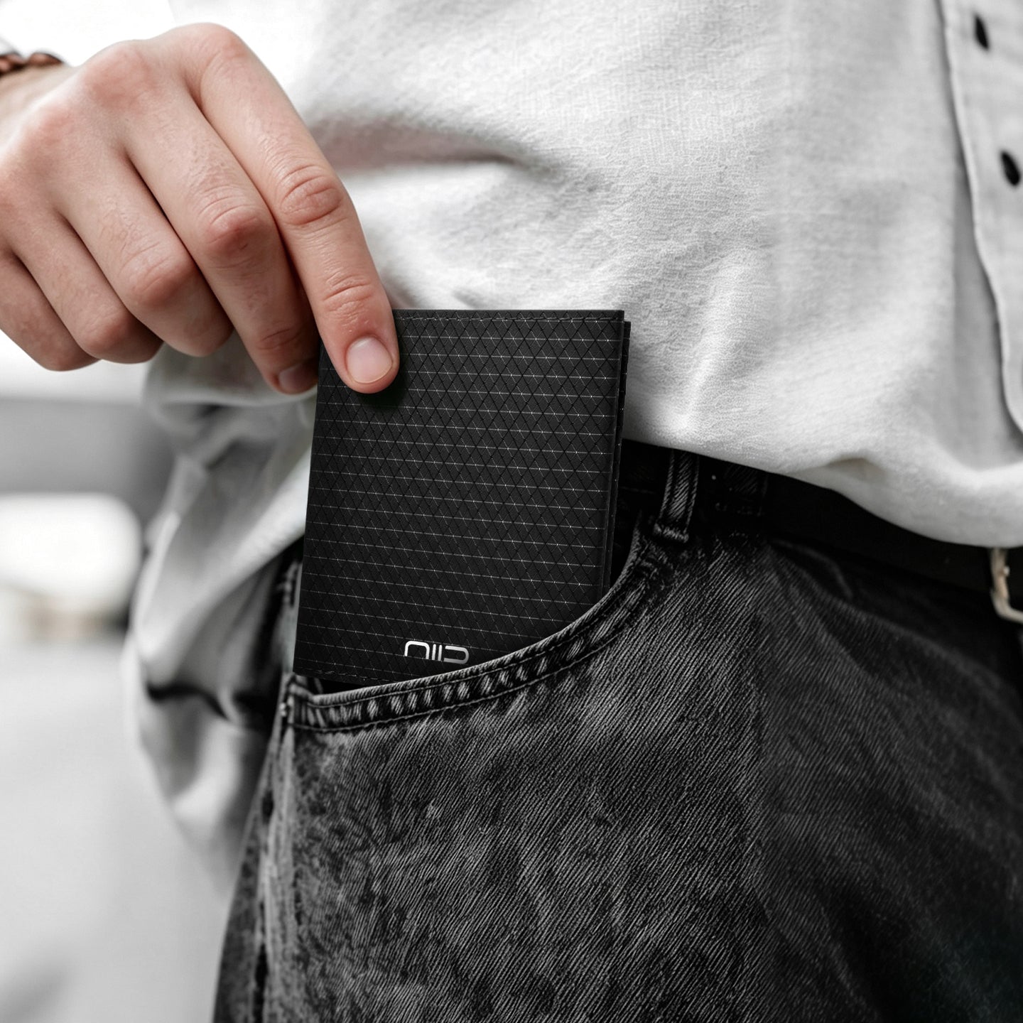 Lifestyle close-up shot of a hand sliding black triangular grid Cordura fabric NIID NEX slim bifold wallet into front jeans pocket; model wears white button shirt and black denim, silver embossed NIID logo visible on wallet, real-world daily carry minimalist EDC scene