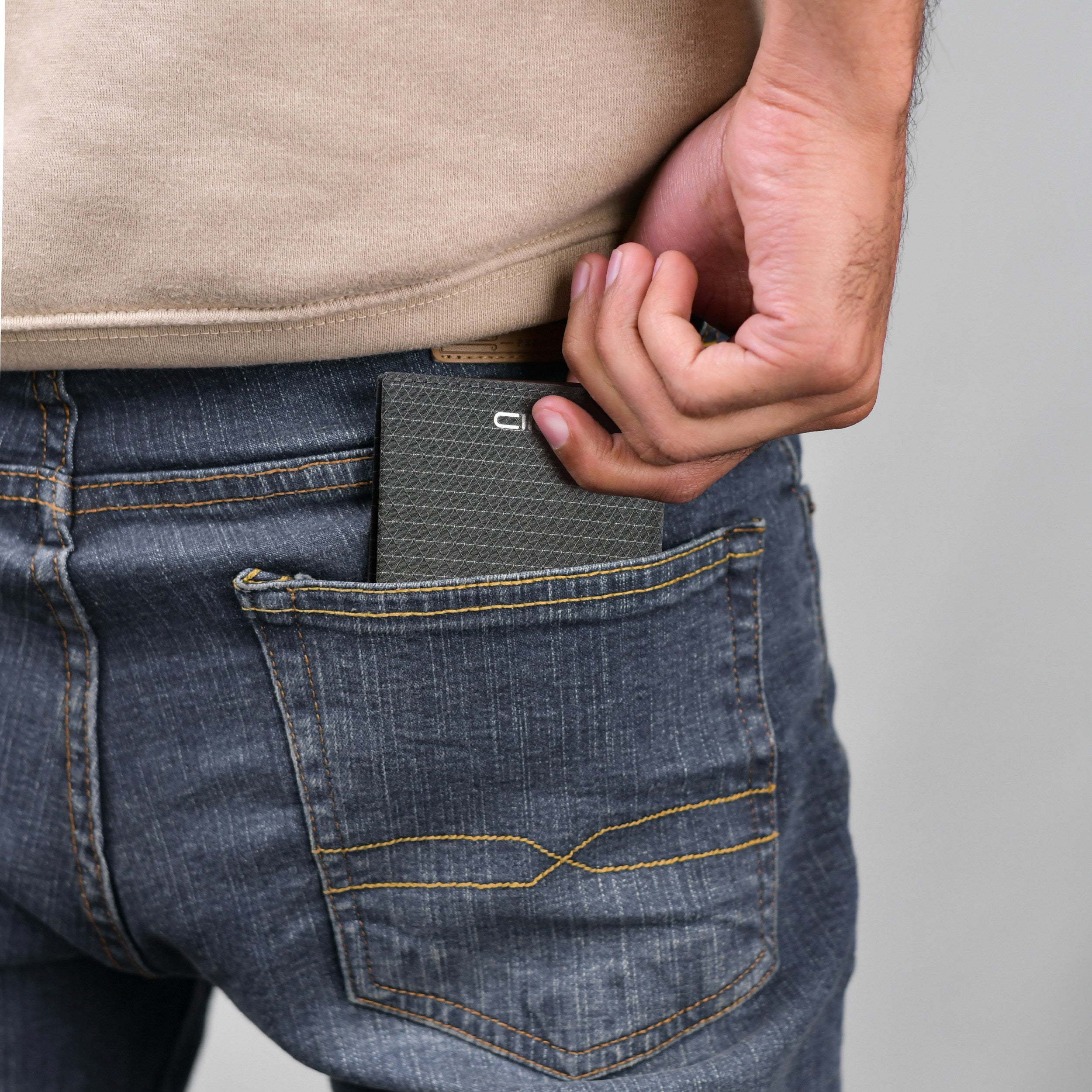 Lifestyle rear pocket shot: a man’s hand pulling dark gray triangular grid Cordura fabric NIID NEX slim bifold wallet out of blue denim jeans back pocket, wearing beige casual sweatshirt, white NIID logo visible on wallet, real daily outdoor carry minimalist EDC scene