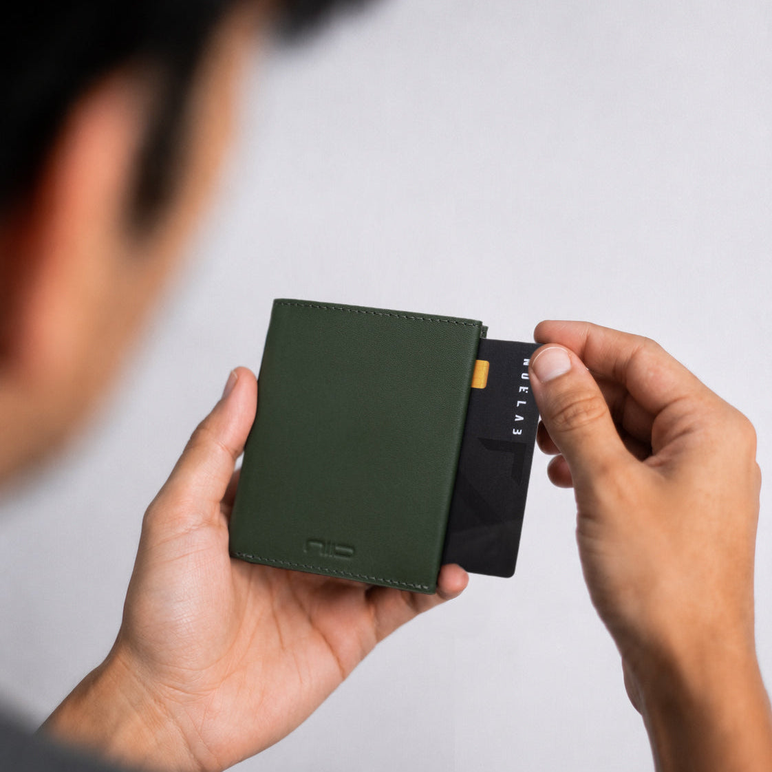 Lifestyle close-up shot of two hands; one holds smooth dark green genuine leather NIID NEX slim bifold wallet with embossed NIID logo, the other slides a black credit card out of the card slot, soft blurred light gray background, minimalist men’s daily carry wallet demonstration photo