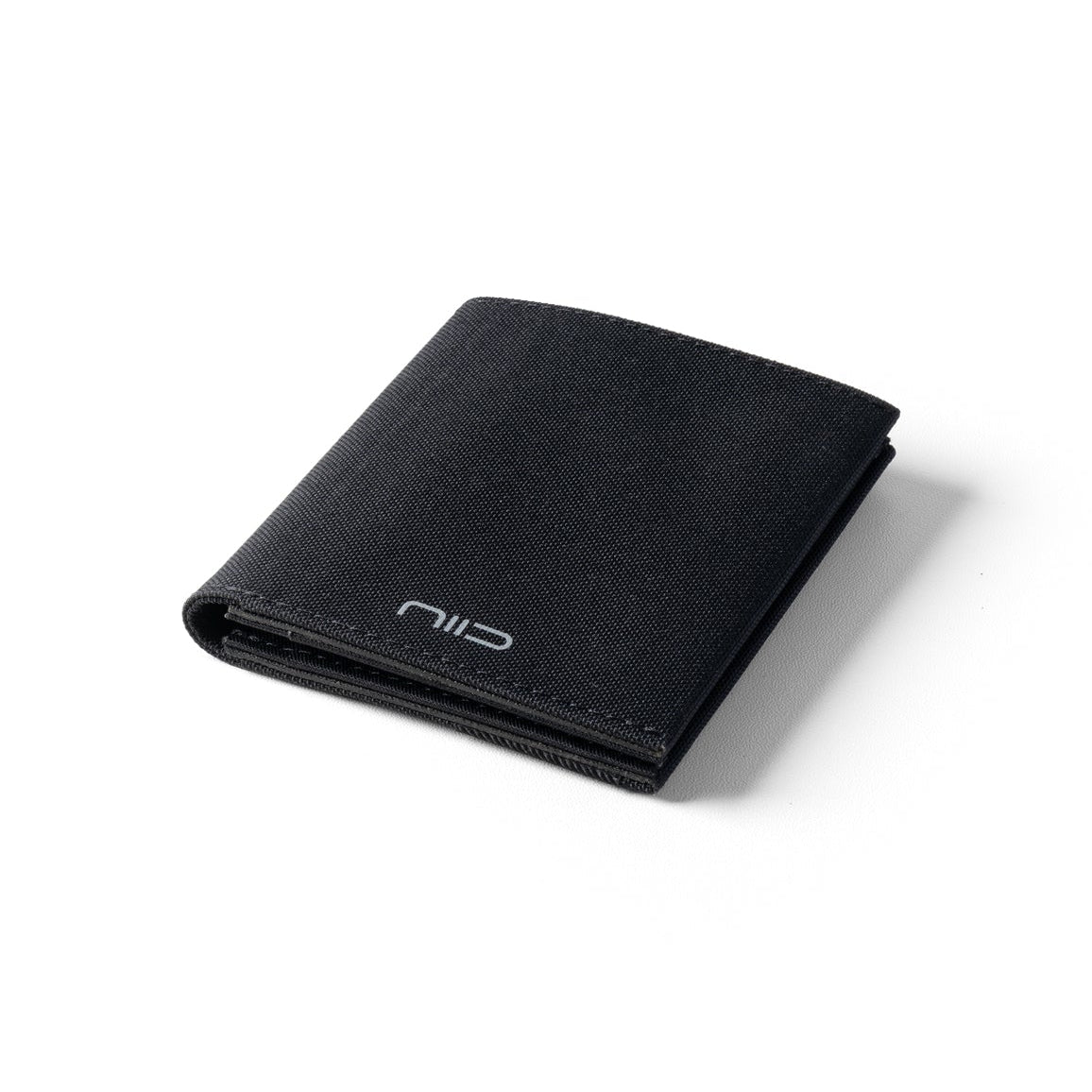 Angled studio shot of solid black Cordura fabric NIID NEX slim bifold wallet resting on clean plain white background, silver embossed NIID logo on lower front, neat edge stitching, soft subtle shadow, minimalist wear-resistant men’s front pocket EDC card holder product photo