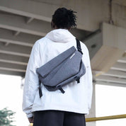 Back view streetwear shot of man in white hooded jacket carrying NIID R1 Radiant Mega grey static crossbody sling bag, concrete bridge urban outdoor background, scratch-proof textured fabric unisex daily carry pack