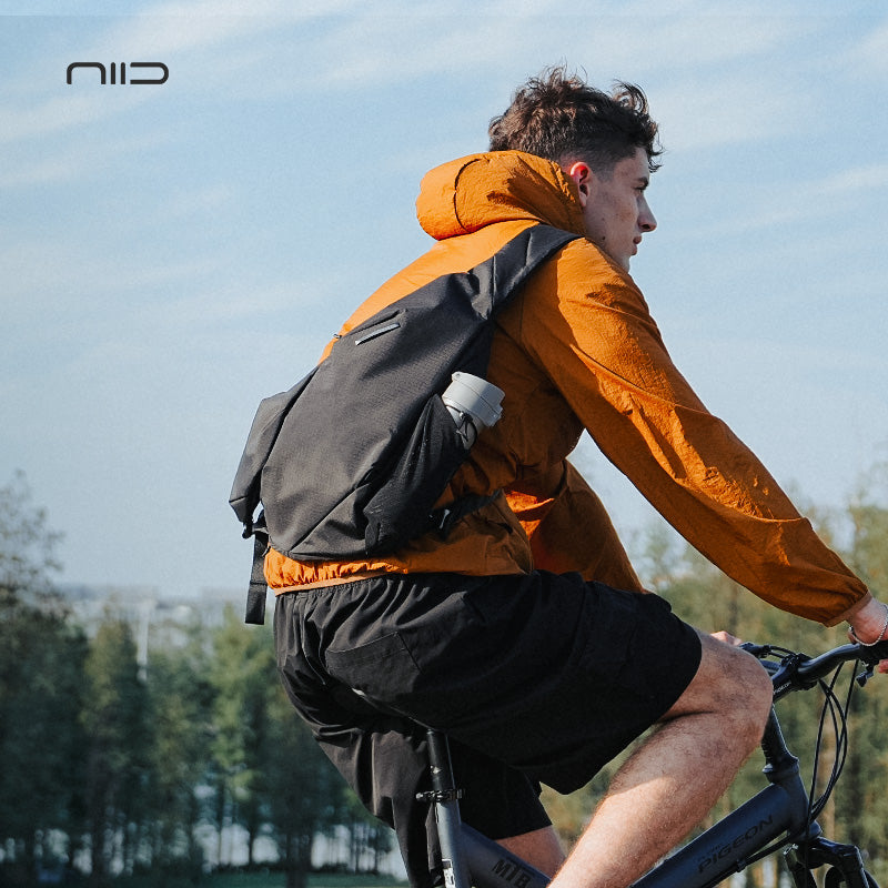 A person riding a bicycle outdoors wearing the NIID R2 Radiant Modular Chest Bag in black, with clear sky and trees in the background.