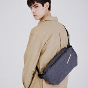 Back view portrait of man in beige jacket carrying NIID RO Plus meteorite black sling crossbody bag with leather brand tag, adjustable black shoulder strap, minimalist light gray studio backdrop for urban daily commute