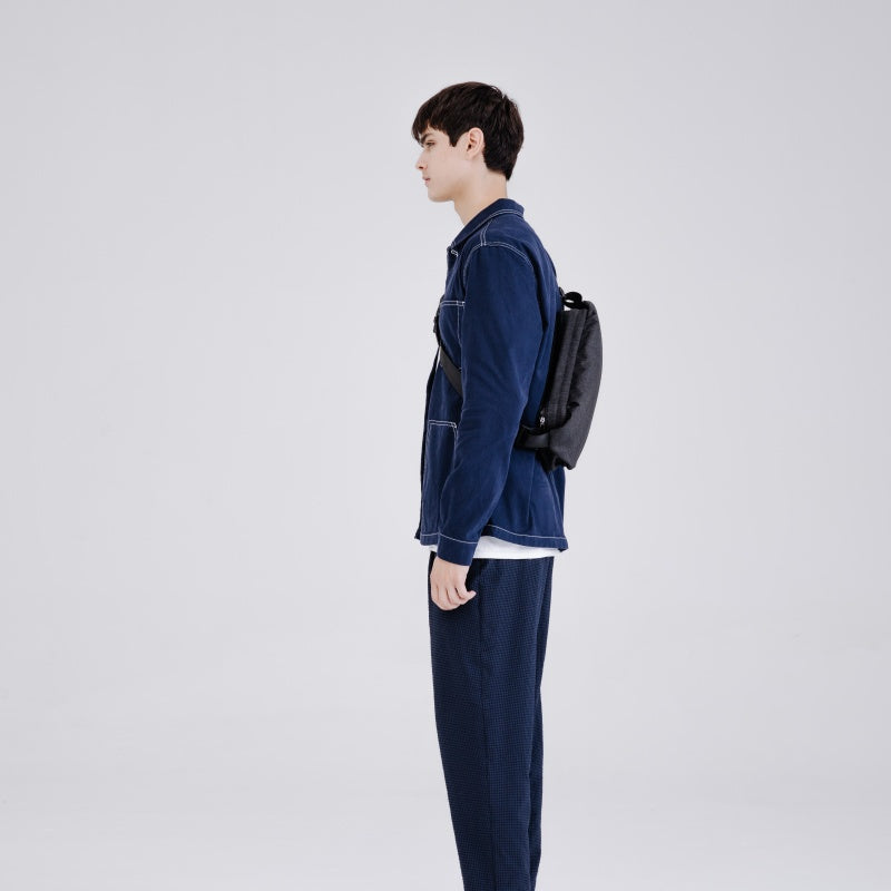 Side profile shot of man in dark denim suit carrying NIID RO Plus meteorite black crossbody sling bag, minimalist urban commute chest pack with adjustable strap, plain light gray studio background