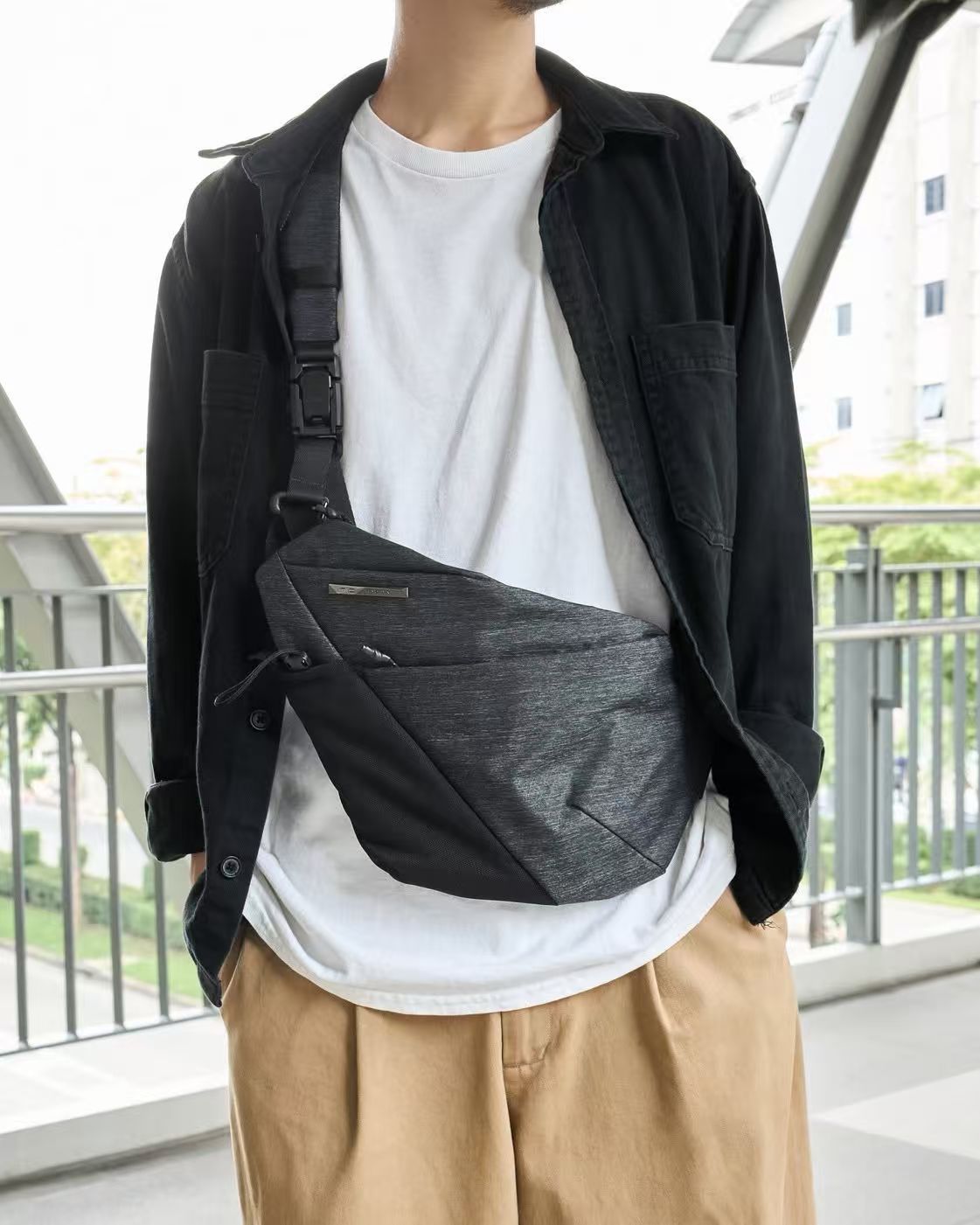 Male model layered with white tee, black oversized shirt and khaki casual pants, wearing NIID Radiant R2 textured dark gray crossbody sling bag with silver metal brand plaque and quick-release buckle strap, outdoor balcony with metal railings and green foliage background, minimalist urban men’s everyday carry fashion photo, soft natural light, no watermarks
