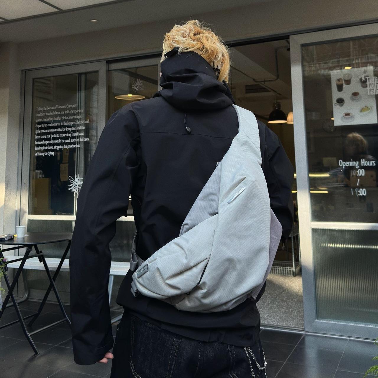 Real‑customer street lifestyle shot of NIID Radiant R2 modular everyday sling bag in Diamond Grid Gray, worn cross‑back outside urban café storefront. Showcases relaxed real‑world carry silhouette for city commute and casual daily outings.